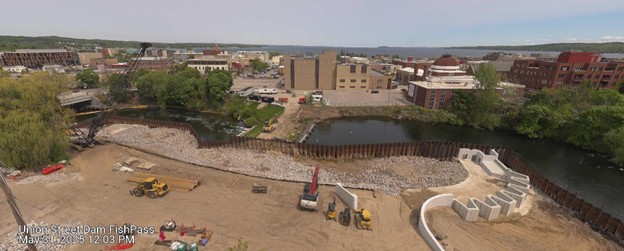 Aerial view of the Union Street Dam FishPass construction site taken on May 21, 2025, at 12:03 PM. The image shows major progress with large sections of riprap placed along the riverbanks and curved concrete structures forming part of the fish passage system. Construction vehicles and equipment are actively staged throughout the site. Sheet piling continues to segment parts of the river. The surrounding downtown area, trees in full foliage, and the bay are visible under a partly cloudy sky.