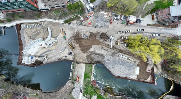 Overhead view of the Union Street Dam FishPass construction site, showing the full width of the river and surrounding area. The river is partially blocked by sheet piling, with active construction on both sides. Curved concrete structures for the fish passage are visible on the left, while large piles of riprap and excavated earth are being shaped on the right. Construction vehicles, materials, and personnel are scattered throughout the site. Trees with spring foliage and adjacent urban buildings border the work zone.