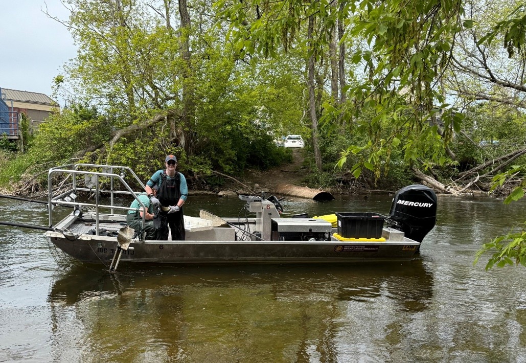 Two people wearing waders and gloves stand on a small aluminum research boat equipped with gear and a Mercury outboard motor. The boat is in a shallow, tree-lined river near a boat launch area, with a white vehicle and drainage pipe visible on the wooded shoreline in the background.